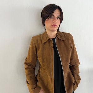 May include: A brown suede jacket with a collar and a zip fastening. The jacket is worn over a black top and blue jeans. The jacket has long sleeves and a tailored fit.