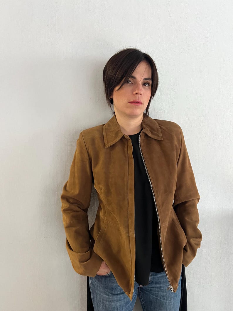 May include: A brown suede jacket with a collar and a zip fastening. The jacket is worn over a black top and blue jeans. The jacket has long sleeves and a tailored fit.