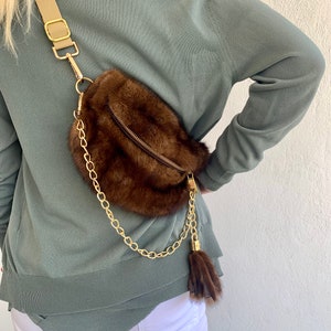 May include: A brown fur crossbody bag with a gold chain strap and a brown fur tassel. The bag has a zipper closure and a small pocket on the front.