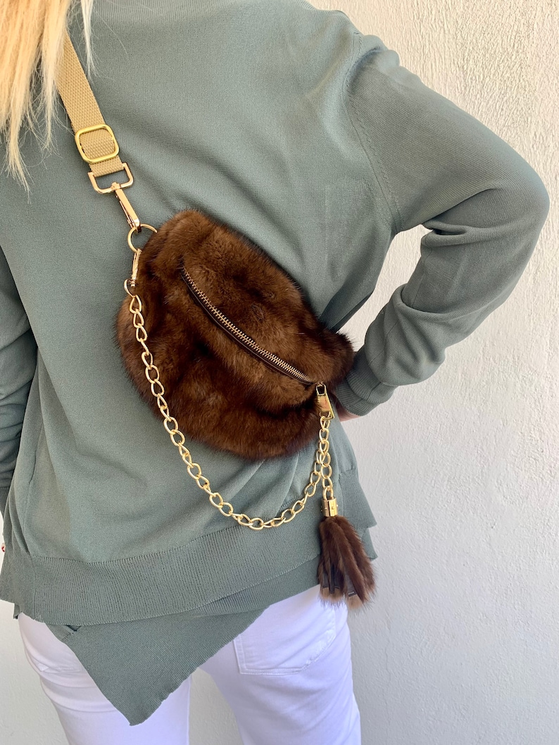 May include: A brown fur crossbody bag with a gold chain strap and a brown fur tassel. The bag has a zipper closure and a small pocket on the front.