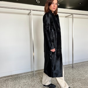 May include: A long, black coat with a textured surface. The coat is worn over light-colored pants and black shoes with silver studs. The coat has a classic, tailored design.