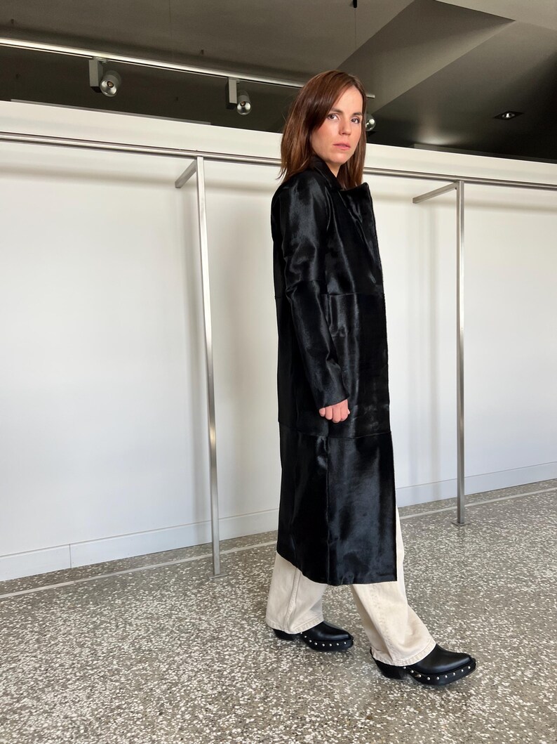 May include: A long, black coat with a textured surface. The coat is worn over light-colored pants and black shoes with silver studs. The coat has a classic, tailored design.