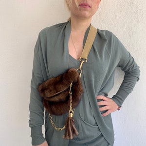 May include: A brown fur crossbody bag with a gold chain and tassel. The bag is worn over a green cardigan.