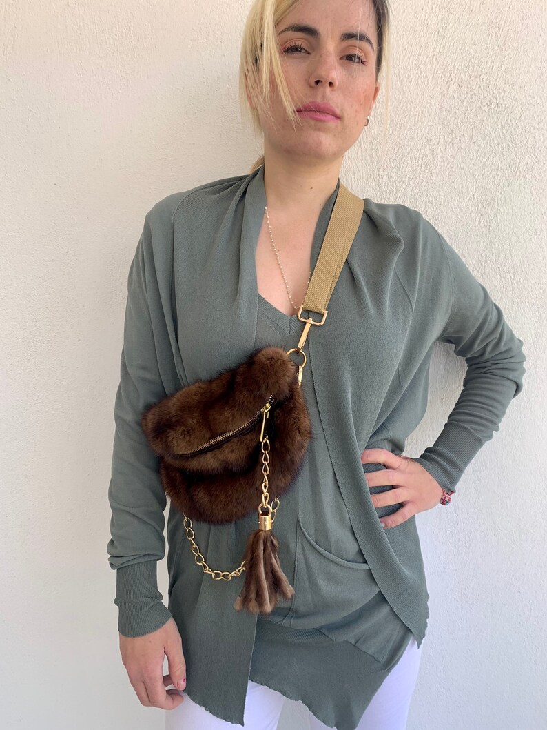 May include: A brown fur crossbody bag with a gold chain and tassel. The bag is worn over a green cardigan.