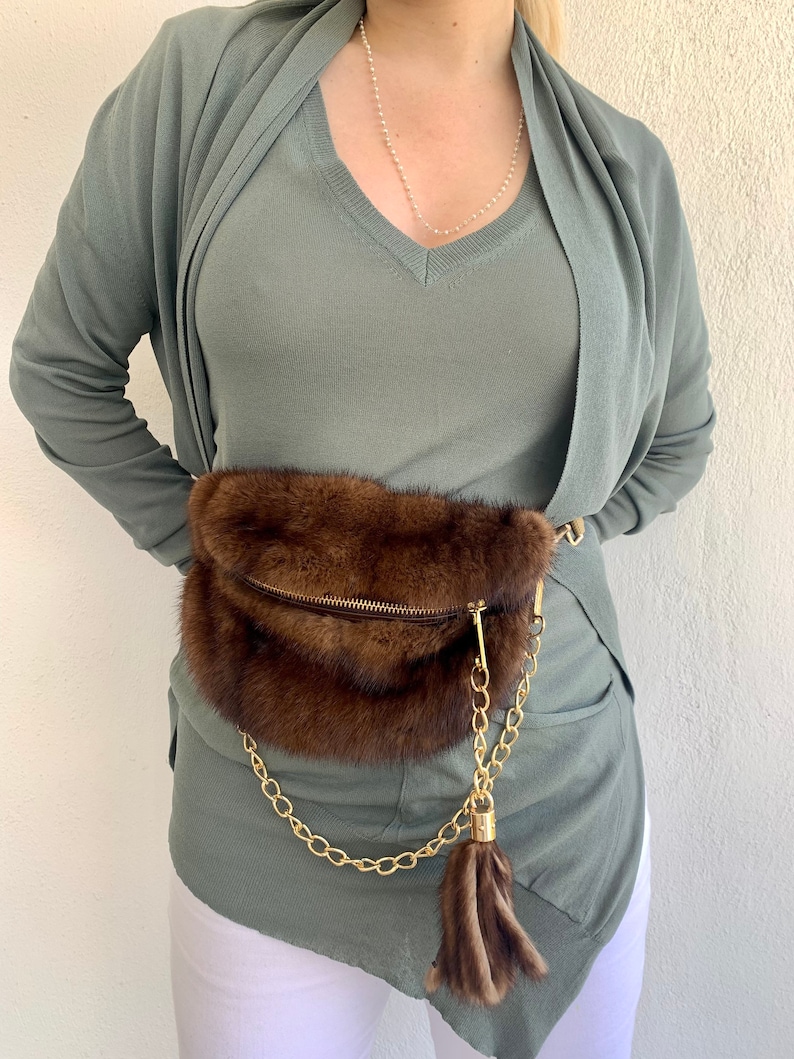 May include: A brown fur waist bag with a gold chain and tassel. The bag is worn over a sage green v-neck top and a matching cardigan.