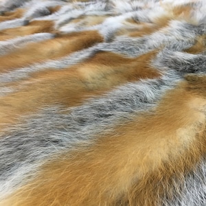 Pode incluir: Um close-up de um tapete de pele macio e felpudo com uma mistura de pele cinza, branca e laranja. A pele est&aacute; disposta em um padr&atilde;o ondulado, criando um visual luxuoso e texturizado.