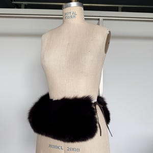 May include: A dark brown fur belt is wrapped around a mannequin. The belt is made of thick, plush fur and is tied with a thin, dark brown leather cord. The mannequin is a light beige color.