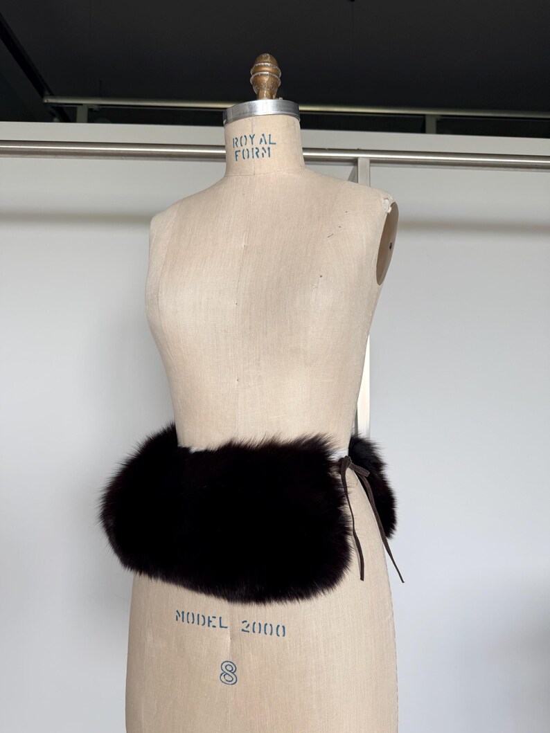 May include: A dark brown fur belt is wrapped around a mannequin. The belt is made of thick, plush fur and is tied with a thin, dark brown leather cord. The mannequin is a light beige color.