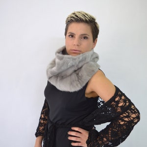 May include: A woman with short blonde hair is wearing a grey fur scarf and a black crochet cardigan. She is looking at the camera.