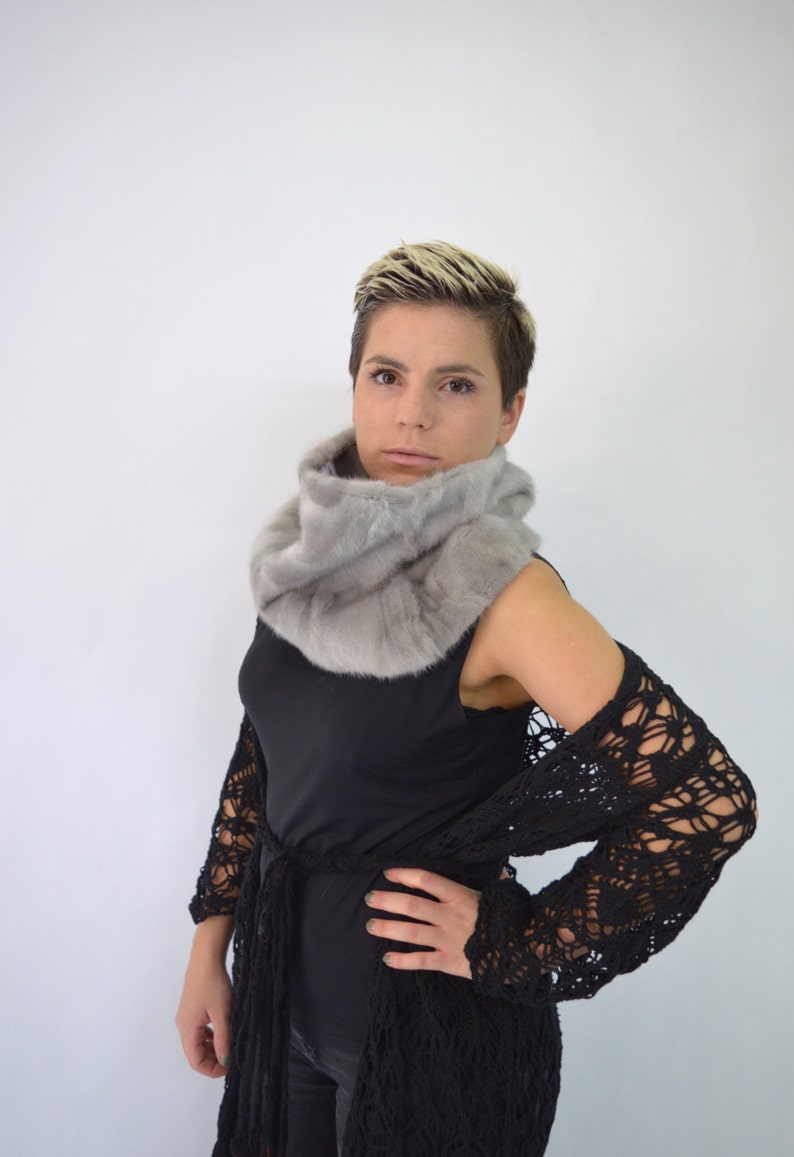 May include: A woman with short blonde hair is wearing a grey fur scarf and a black crochet cardigan. She is looking at the camera.
