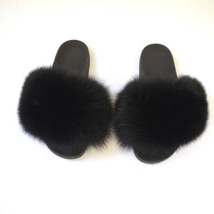 K&ouml;nnte beinhalten: Ein Paar schwarze, pelzgef&uuml;tterte Hausschuhe. Die Hausschuhe haben einen schwarzen Pelzbesatz an den R&auml;ndern und eine schwarze Sohle.