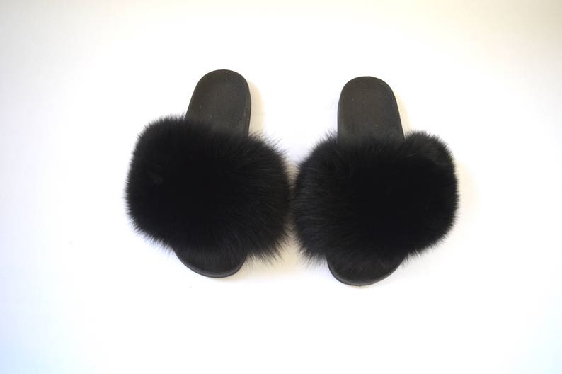 K&ouml;nnte beinhalten: Ein Paar schwarze, pelzgef&uuml;tterte Hausschuhe. Die Hausschuhe haben einen schwarzen Pelzbesatz an den R&auml;ndern und eine schwarze Sohle.