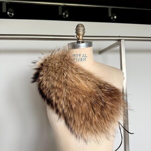 Op de afbeelding: Een bruine bontkraag met zwarte uiteinden, gedrapeerd over een mannequin. Het bont ziet er zacht en pluizig uit, met een mix van lichte en donkere bruintinten. De mannequin heeft de tekst "ROYAL FORM".
