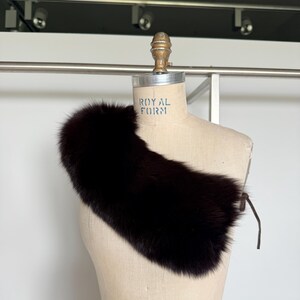 May include: A dark brown fur collar with a leather tie closure. The collar is displayed on a mannequin with the text "ROYAL FORM". The fur appears soft and plush, with a rich, dark color.
