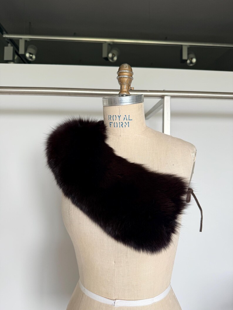 May include: A dark brown fur collar with a leather tie closure. The collar is displayed on a mannequin with the text "ROYAL FORM". The fur appears soft and plush, with a rich, dark color.