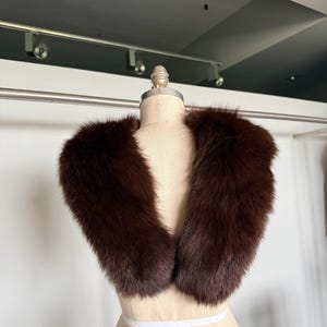 May include: A dark brown fur collar displayed on a mannequin. The collar is a rich, deep brown color and appears soft and plush. The fur is thick and evenly distributed, creating a luxurious appearance. The collar is designed to be worn around the neck.