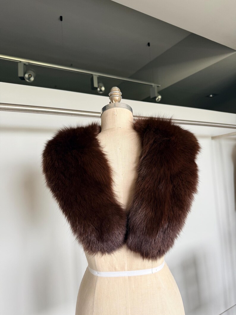 May include: A dark brown fur collar displayed on a mannequin. The collar is a rich, deep brown color and appears soft and plush. The fur is thick and evenly distributed, creating a luxurious appearance. The collar is designed to be worn around the neck.