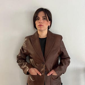 May include: A brown leather blazer with a notched collar, two front pockets, and button fastenings. The blazer is worn over a black top and blue jeans. The model is standing in front of a white wall.