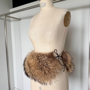Op de afbeelding: Een bruine en zwarte bontkraag met een zwarte striksluiting, getoond op een mannequin. Het bont heeft licht- en donkerbruine tinten, met zwarte punten. De mannequin is beige en staat voor een witte muur.