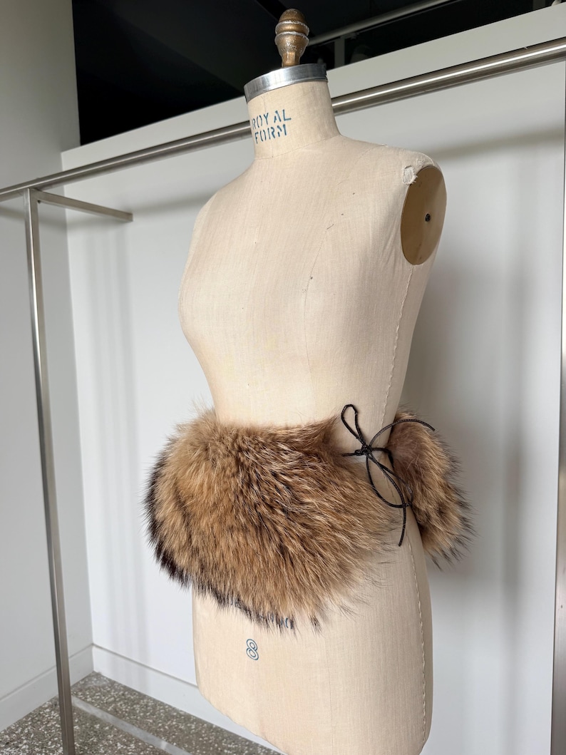 Op de afbeelding: Een bruine en zwarte bontkraag met een zwarte striksluiting, getoond op een mannequin. Het bont heeft licht- en donkerbruine tinten, met zwarte punten. De mannequin is beige en staat voor een witte muur.