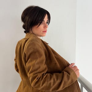 May include: A tan suede jacket with a traditional collar and button fastening. The jacket has a fitted shape and is worn open, showing the inside lining. The jacket is a warm, earthy colour, suitable for various occasions.