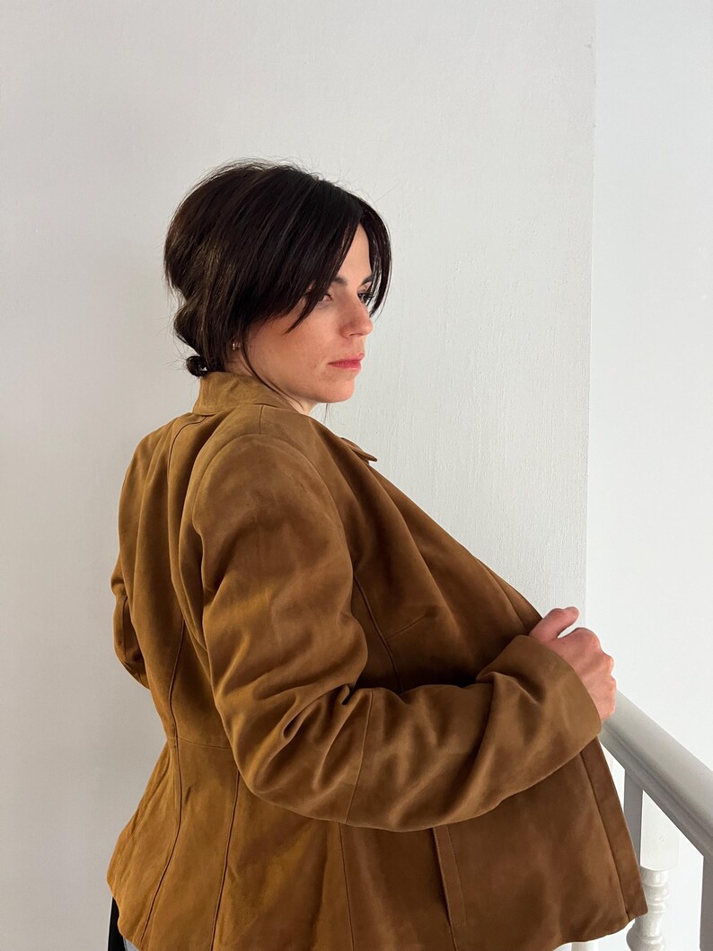 May include: A tan suede jacket with a traditional collar and button fastening. The jacket has a fitted shape and is worn open, showing the inside lining. The jacket is a warm, earthy colour, suitable for various occasions.