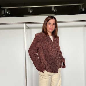 Può includere: Un blazer marrone con un motivo a foglie. Il blazer ha maniche lunghe e un colletto classico. La modella indossa un top color crema e pantaloni chiari. Un capo d'abbigliamento elegante.