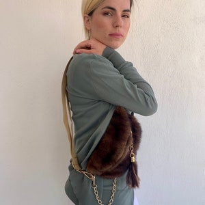 May include: A woman wearing a green long-sleeved shirt and white pants is wearing a brown fur fanny pack with a gold chain and tassel.