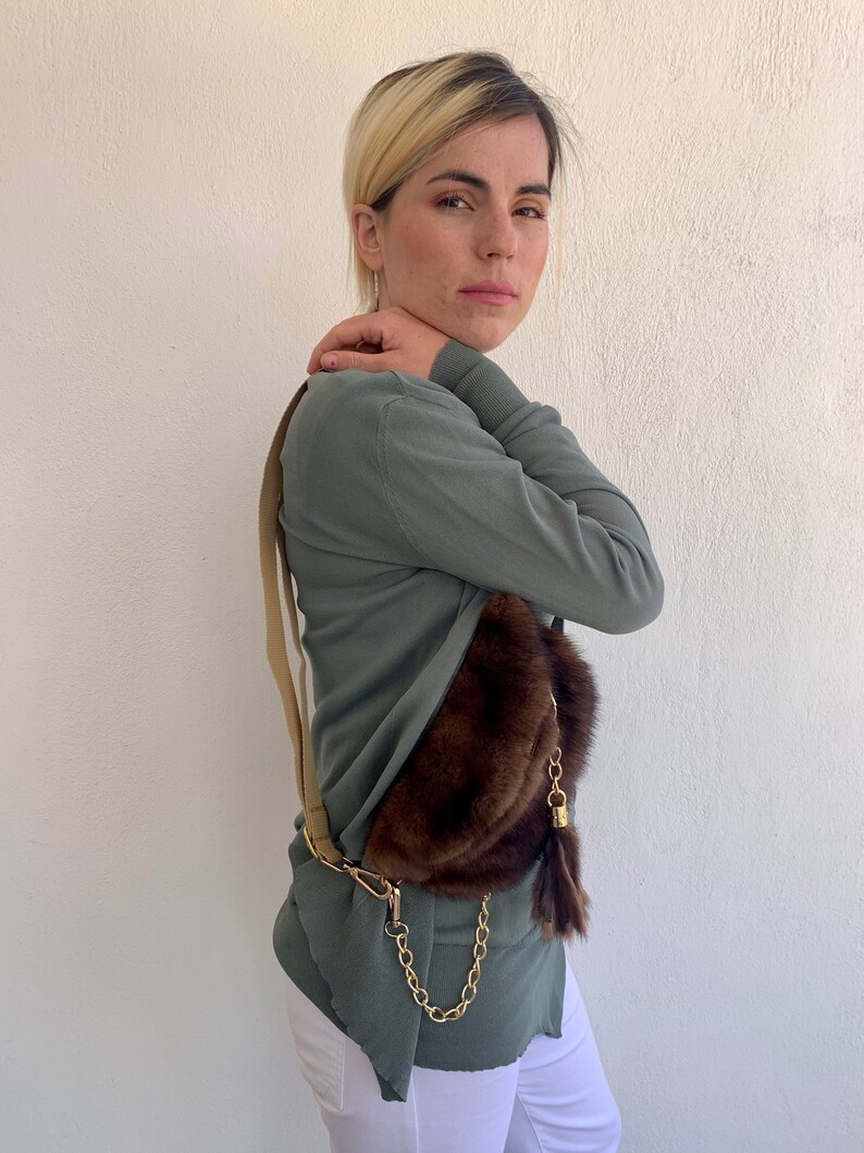 May include: A woman wearing a green long-sleeved shirt and white pants is wearing a brown fur fanny pack with a gold chain and tassel.