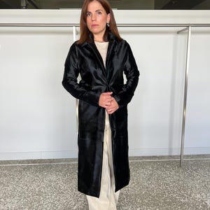 May include: A long, black coat with a velvet-like texture. The coat has a classic lapel collar and a tailored fit. The model is wearing a cream-colored top and light-colored pants. The coat appears to be knee-length.