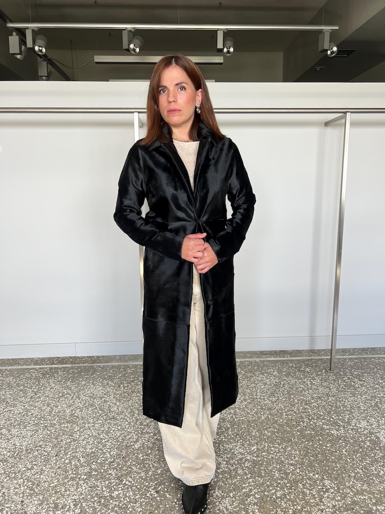 May include: A long, black coat with a velvet-like texture. The coat has a classic lapel collar and a tailored fit. The model is wearing a cream-colored top and light-colored pants. The coat appears to be knee-length.