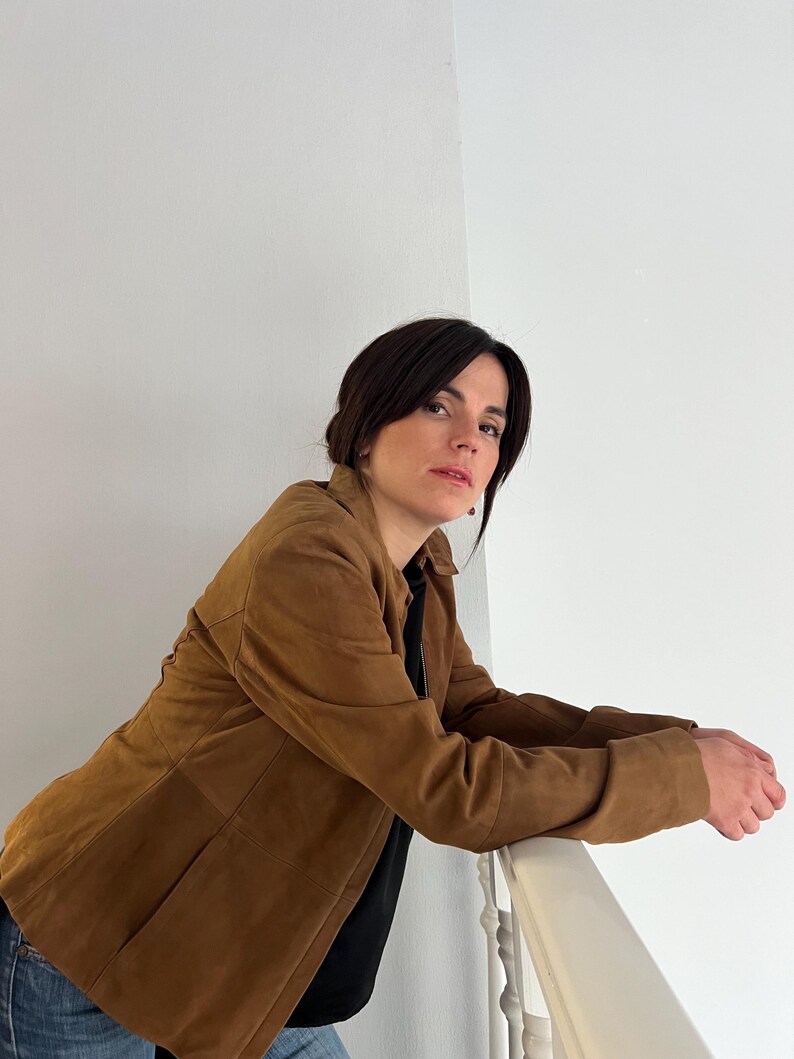 May include: A tan suede jacket with a tailored fit. The jacket features a classic collar, long sleeves, and a button closure. The person is wearing a black top and blue jeans.