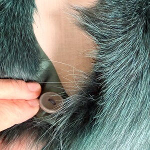 K&ouml;nnte beinhalten: Nahaufnahme eines teal-farbenen Pelzkragens mit Knopfverschluss. Das Fell hat eine Mischung aus Teal- und Silbert&ouml;nen, wodurch ein strukturiertes Aussehen entsteht. Eine Hand wird gezeigt, die den Knopf befestigt, der eine ged&auml;mpfte braune Farbe hat.