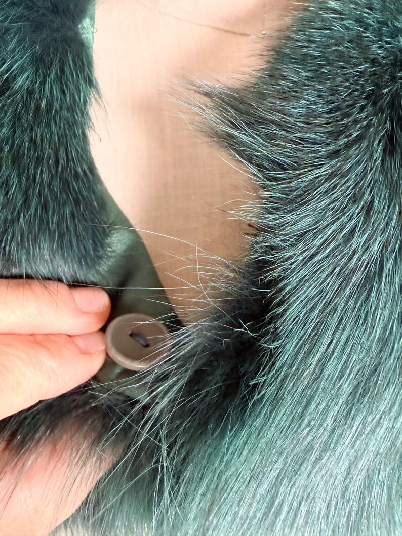 K&ouml;nnte beinhalten: Nahaufnahme eines teal-farbenen Pelzkragens mit Knopfverschluss. Das Fell hat eine Mischung aus Teal- und Silbert&ouml;nen, wodurch ein strukturiertes Aussehen entsteht. Eine Hand wird gezeigt, die den Knopf befestigt, der eine ged&auml;mpfte braune Farbe hat.
