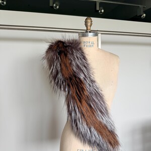 May include: A luxurious fur stole with a mix of brown, silver, and reddish-brown tones. The stole is draped over a mannequin, showcasing its texture and design. The fur appears soft and fluffy, perfect for adding a touch of elegance.
