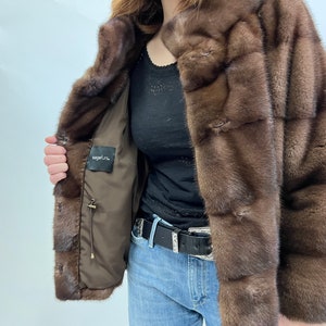 K&ouml;nnte beinhalten: Eine braune Pelzjacke mit braunem Futter und einem Etikett mit der Aufschrift "sagafurs". Die Jacke hat einen Kragen und einen Knopfverschluss.