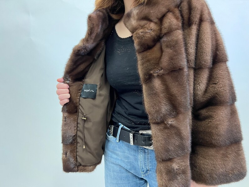 K&ouml;nnte beinhalten: Eine braune Pelzjacke mit braunem Futter und einem Etikett mit der Aufschrift "sagafurs". Die Jacke hat einen Kragen und einen Knopfverschluss.