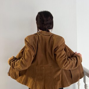 May include: A brown suede jacket with a classic collar and long sleeves. The jacket is open, revealing the interior lining. The back view shows the tailored fit and the texture of the suede material. The jacket is part of a clothing collection.