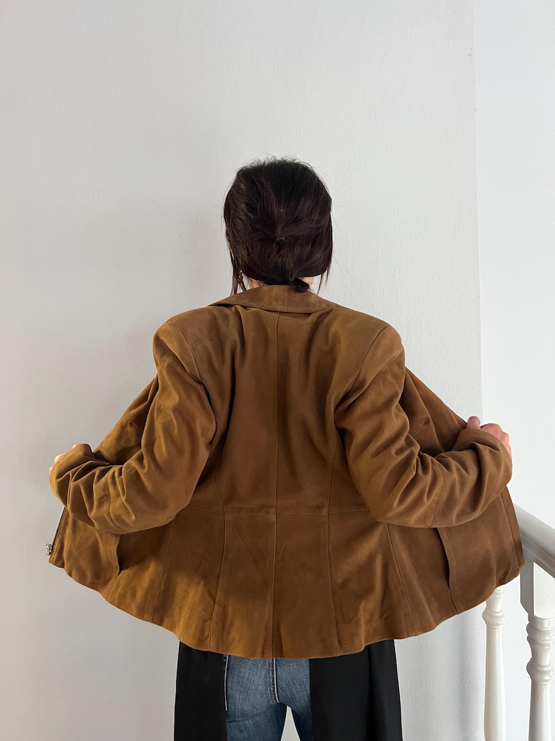 May include: A brown suede jacket with a classic collar and long sleeves. The jacket is open, revealing the interior lining. The back view shows the tailored fit and the texture of the suede material. The jacket is part of a clothing collection.