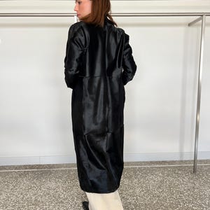 May include: A long, black coat with a shiny, textured appearance. The coat is knee-length and has a tailored fit. The person is wearing cream-colored pants and black shoes.