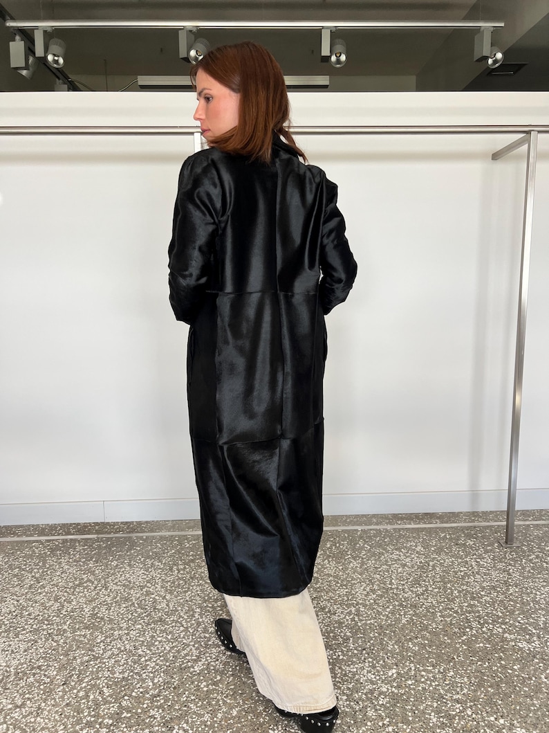 May include: A long, black coat with a shiny, textured appearance. The coat is knee-length and has a tailored fit. The person is wearing cream-colored pants and black shoes.