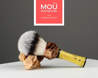 May include: A shaving brush with a black and white badger hair brush head and a yellow and brown wooden handle. The handle has a black band around it. The brush is sitting on a light brown rock.
