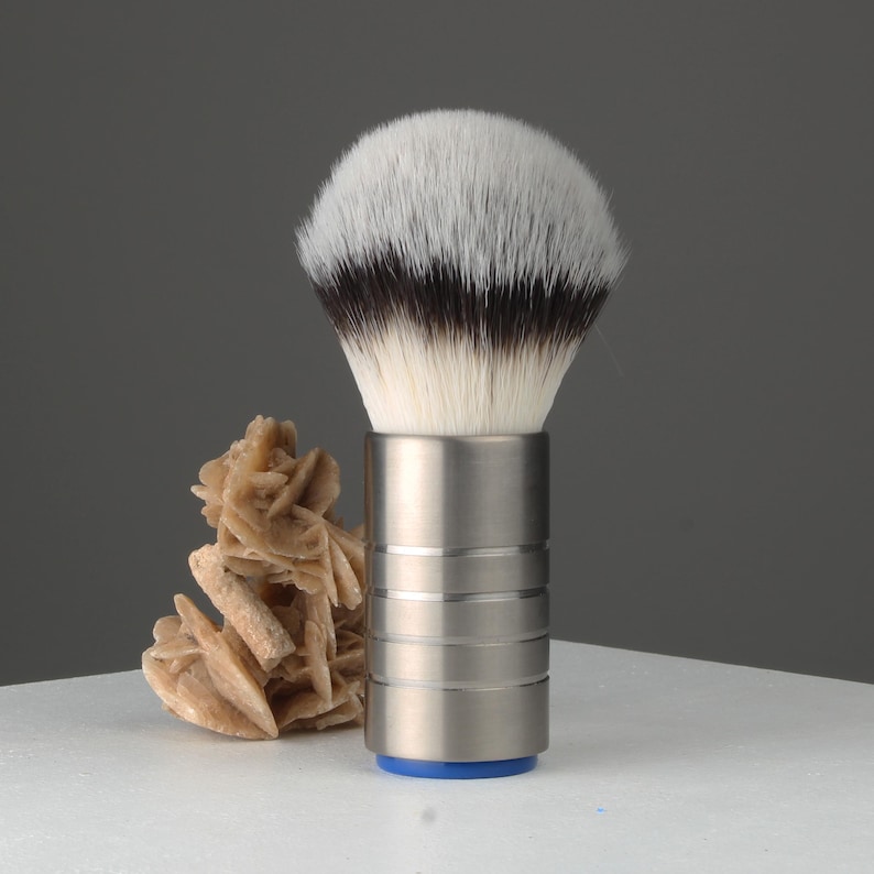 Op de afbeelding: Een zilveren scheerkwast met een wit en zwart borstelhoofd van dassenhaar. De kwast heeft een zilveren handvat met een blauwe basis.