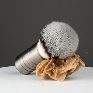 Op de afbeelding: Een zilveren en blauwe scheerkwast met een wit en zwart borstelhoofd van dassenhaar. De kwast rust op een lichtbruine steen.