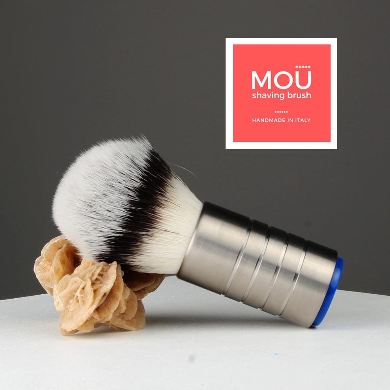 Op de afbeelding: Een zilveren scheerkwast met een blauwe basis en een wit en zwart borstelhoofd van dassenhaar. De kwast ligt op een wit oppervlak met een lichtbruine steen ernaast. De tekst "MOU shaving brush HANDMADE IN ITALY" staat op een rood vierkant.