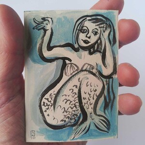 Può includere: Piccolo dipinto rettangolare di una sirena, con contorni a inchiostro nero e lavaggi ad acquerello. La sirena ha lunghi capelli neri, un top a conchiglia e una coda di pesce. Lo sfondo è azzurro.