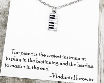 May include: Silver-toned necklace with a piano key pendant. The pendant features black and white keys. The necklace is displayed on a white card with a quote by Vladimir Horowitz about the piano.