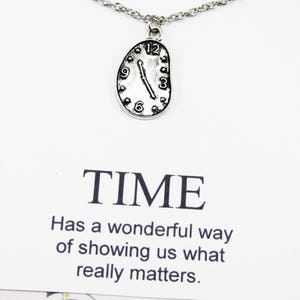 Puede incluir: Un collar de plata con un colgante en forma de reloj derritiéndose. El colgante tiene marcas de números romanos negros. El collar viene con una tarjeta que dice "TIME Has a wonderful way of showing us what really matters."