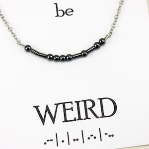 Puede incluir: Un collar de cadena plateada con cuentas negras que deletrea "WEIRD" en braille. El collar está sobre una tarjeta blanca con la palabra "be" y la palabra "WEIRD" impresa en negro.