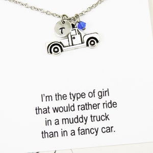 Op de afbeelding: Een zilveren ketting met een hanger in de vorm van een pick-up truck, een blauwe kristal en een zilveren schijf met de letter "T" erop. De ketting wordt geleverd met een kaart waarop staat: "Ik ben het type meisje dat liever in een modderige truck rijdt dan in een chique auto."